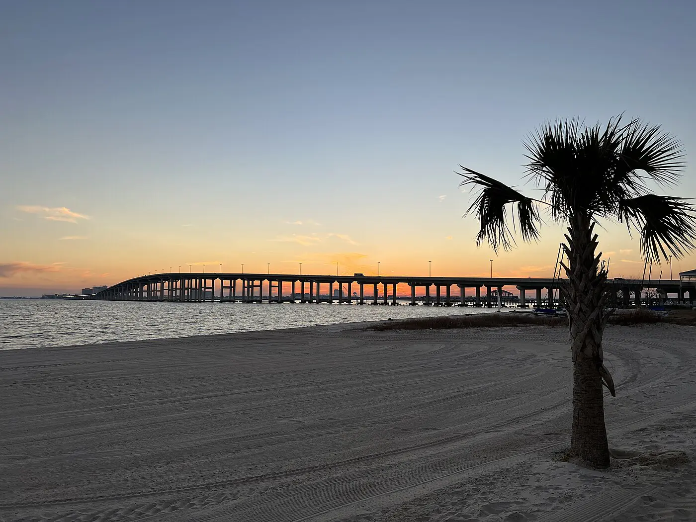 A palm on the sand at Front Beach with the Biloxi Bay Bridge arcing across an orange dusk sky